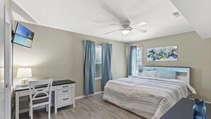 A bedroom with a bed, desk, chair, ceiling fan, and artwork on the wall.