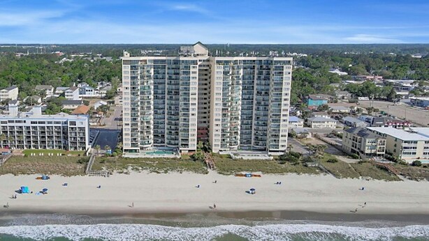 A high-rise hotel with multiple floors, a beach in the foreground, and a cityscape in the background.