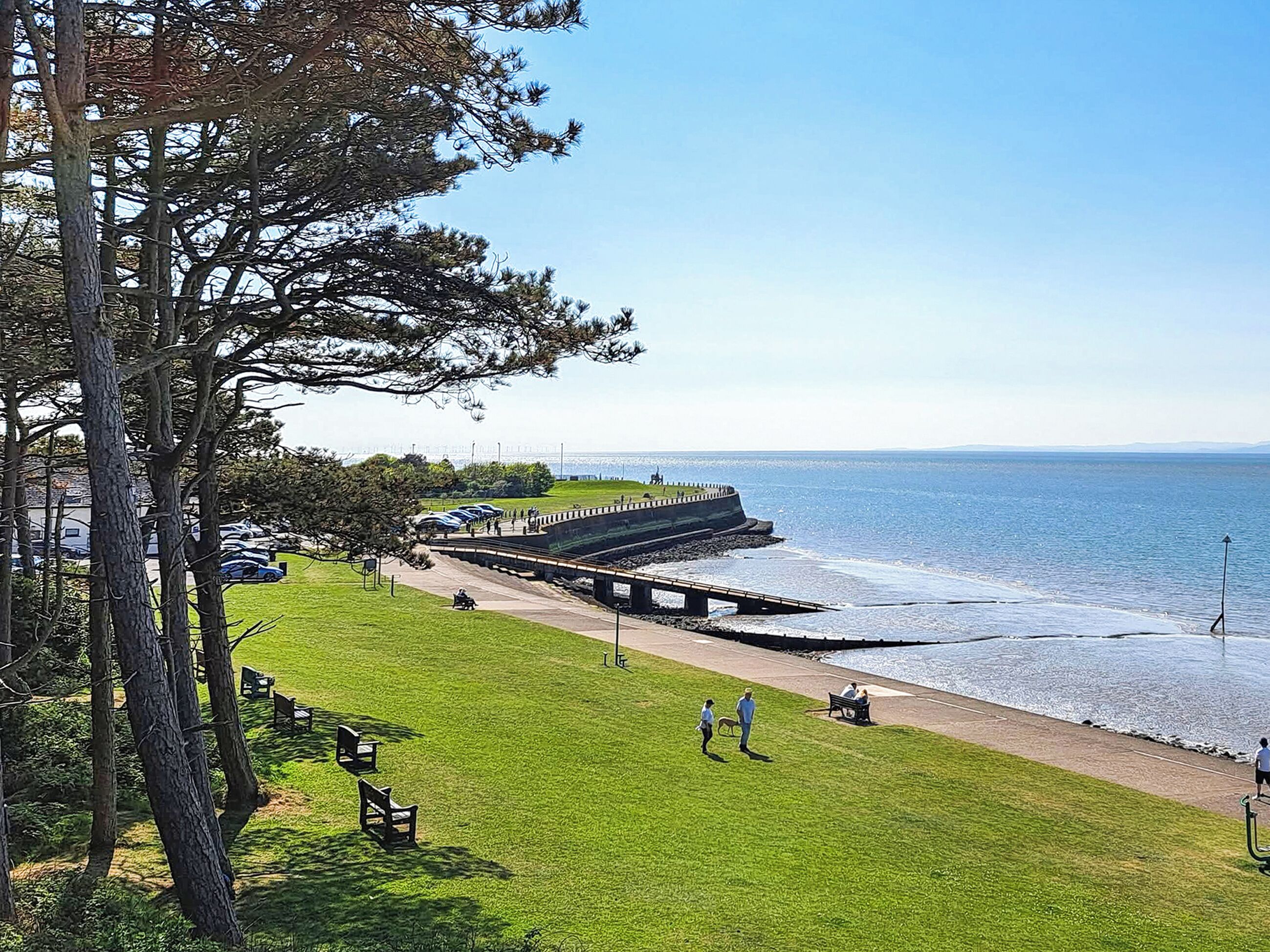 A coastal park with a grassy area, benches, a walking path, and a pier extending into the water.