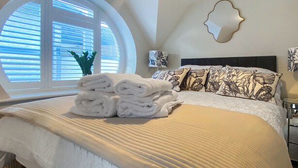 A neatly made bed with rolled white towels, a round window with blinds, and a wall-mounted decorative mirror. - The Police Station - UK45642 (Silloth)