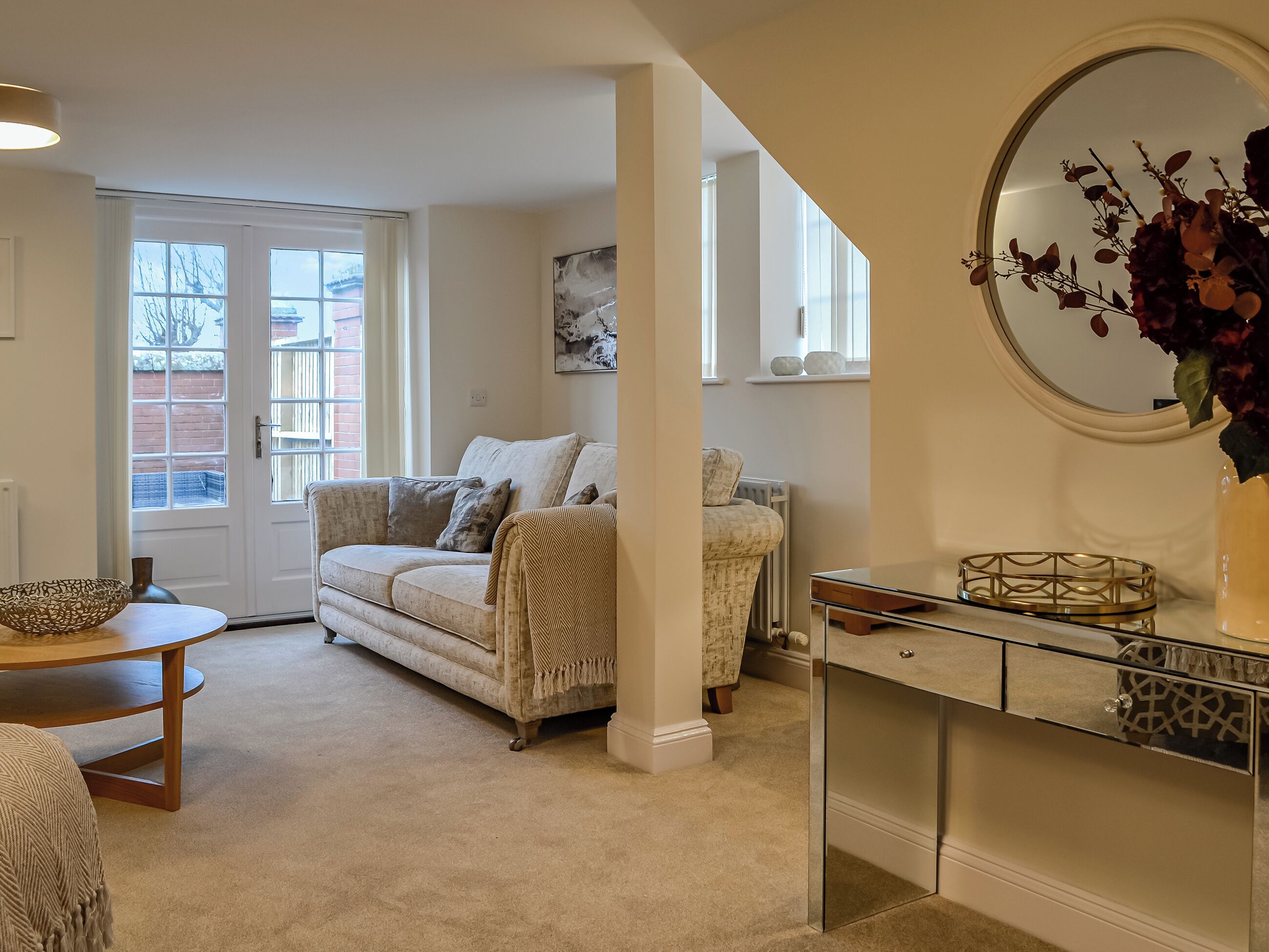 Una sala de estar con un sofá beige, una mesa de centro de cristal y un espejo decorativo.