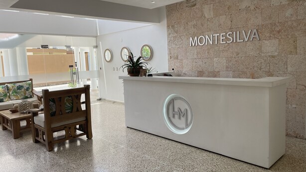 En reception med et skilt, der viser "MONTESILVA", og en logo, i et moderne interiør med en stenvæg, træmøbler og et flisegulv.