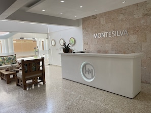 A reception desk with a sign reading "MONTESILVA" and a logo, surrounded by a modern interior with a stone wall, wooden furniture, and a tiled floor. - Hotel Montesilva  (Puerto Plata)