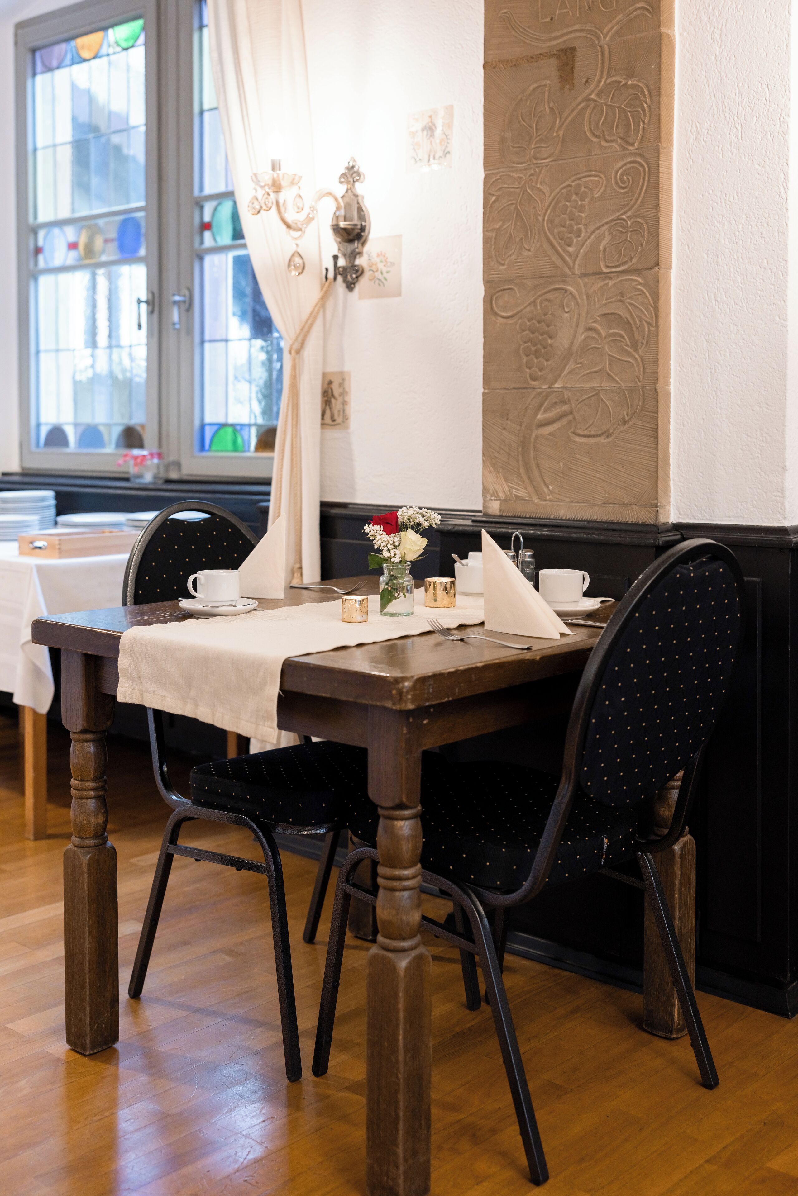 A dining area with a wooden table set for two, black chairs with gold patterns, a white tablecloth, and a vase with flowers.