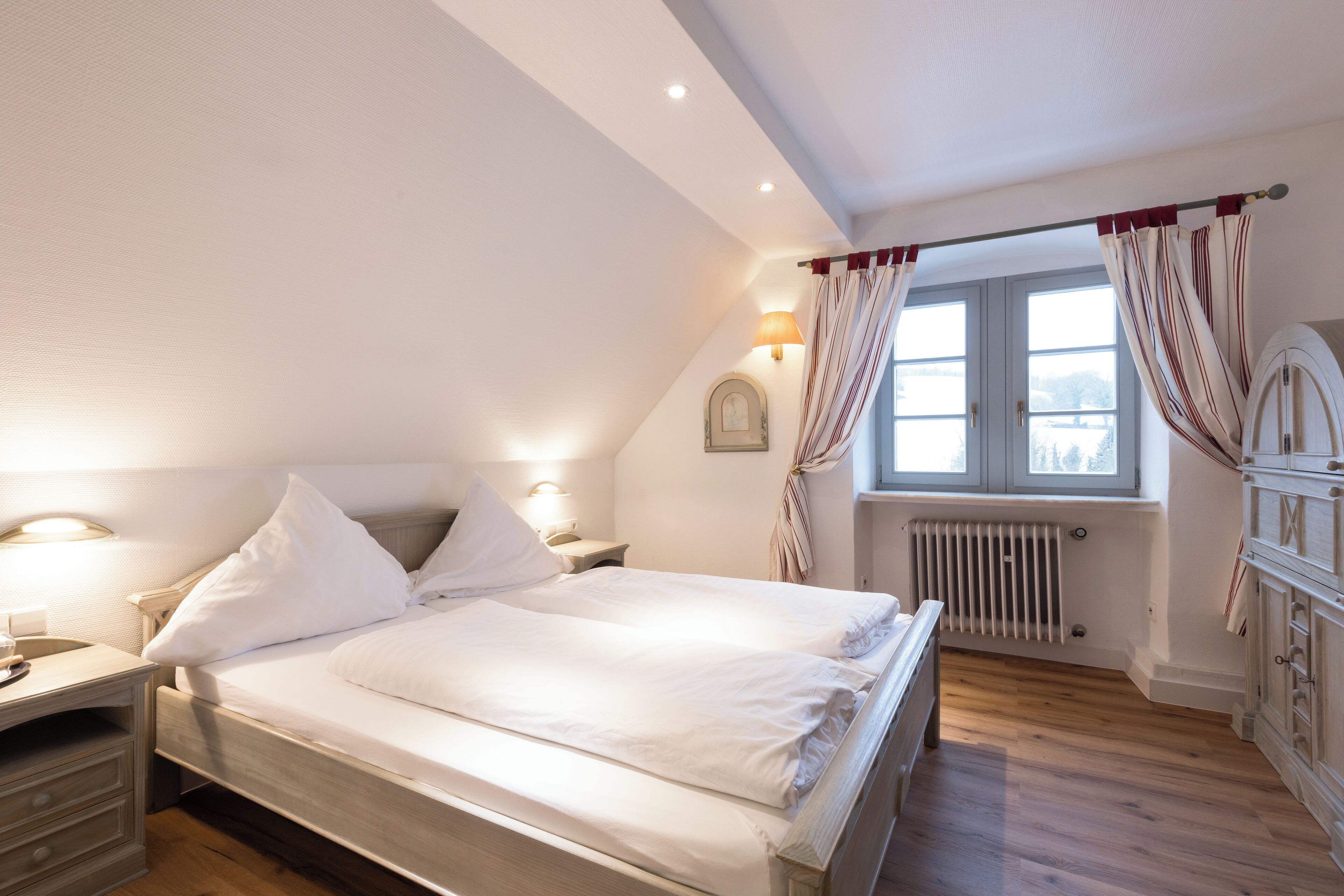 Ein Schlafzimmer mit einem hölzernen Bett, weißer Bettwäsche, einem Nachttisch mit Lampe, einem Fenster mit Vorhängen und einer Heizung.