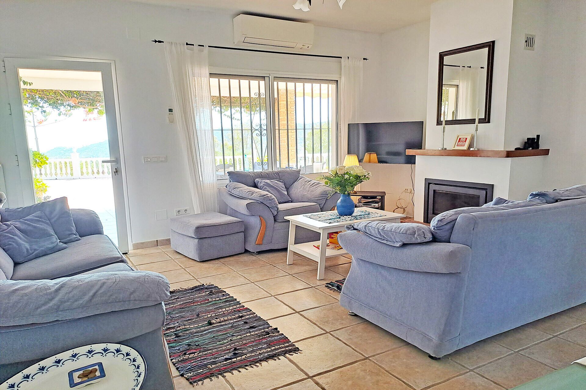 Ein Wohnzimmer mit grauen Sofas, einem kleinen Tisch mit einer Vase, einem Kamin und einem großen Fenster mit Vorhängen.