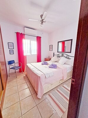 A bedroom with a bed, a chair, a ceiling fan, a window with curtains, and a mirror on the wall.