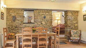A dining area with a stone wall, wooden chairs, a table with plates and a vase of flowers, and a sofa in the background. - Picked for its character and only a 5 minute drive to the beach. (Keeston)
