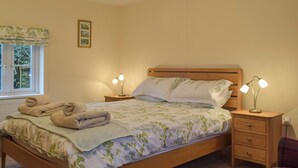 A neatly made bed with a floral bedspread, two bedside lamps, a wooden nightstand, and a window with a view of greenery. - Picked for its character and only a 5 minute drive to the beach. (Keeston)