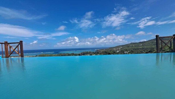En klarblå pool med en träkonstruerad poolbrygga, omgiven av frodig grönska och med utsikt över havet.
