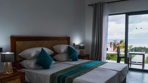 Habitación de hotel con una cama grande, dos lámparas de noche, vista al océano y un balcón con mobiliario exterior.
