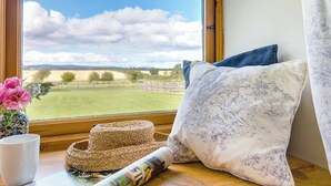 Vista desde una ventana: un campo verde, una cesta tejida, una revista enrollada y un cojín decorativo sobre un repisa de madera.