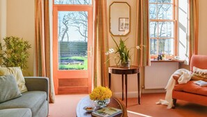 Una sala de estar acogedora con un sofá, una mesa de centro con flores y libros, una mesita auxiliar y una ventana con vistas a árboles.