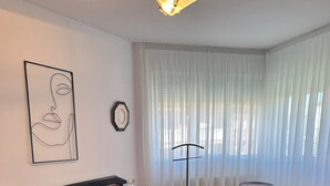 A bedroom with a wooden bed, a bedside table, a floor lamp, and a view through sheer curtains.