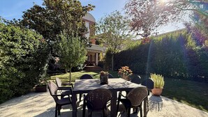 A patio with a table and chairs, surrounded by greenery and a building in the background.