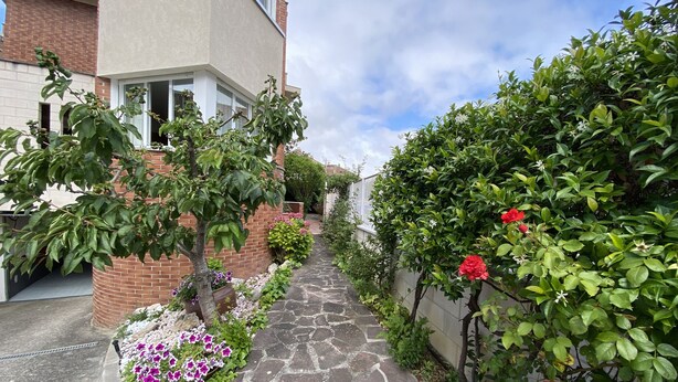 A stone pathway flanked by lush greenery and flowering plants leading to a building with a brick facade and large windows.