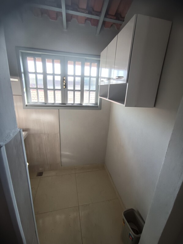A small, enclosed room with a window, tiled floor, and a trash bin. - Jardim das Orquídeas 2 (Rio das Ostras)