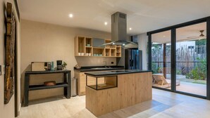 A modern kitchen with a central island, wooden shelves, and a large sliding glass door to a backyard. - Villa Tovar Tulum / Private Pool/ Starlink Wi-Fi / Mayan Jungle.  (Tulum)
