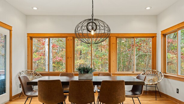 Comedor con mesa de madera, cuatro sillas, lámpara colgante y grandes ventanas con vistas a follaje otoñal.