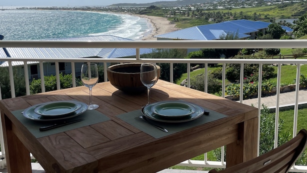 Una mesa de madera con dos copas de vino y platos en un balcón con vista a una playa y al océano.