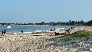 Una playa con gente caminando y sentada, un barco en el agua y edificios al fondo.