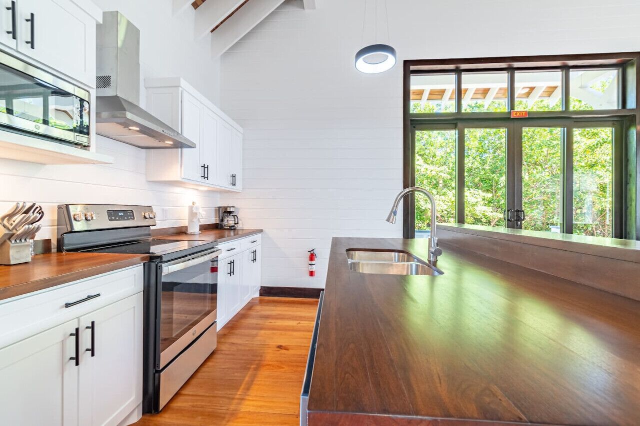 Moderni keittiö, jossa on valkoiset kaapit, ruostumattomasta teräksestä valmistetut kodinkoneet ja suuri ikkuna, josta avautuu näkymä vehreään maisemaan.