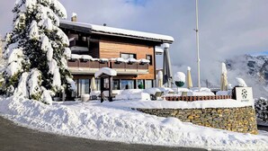 A snow-covered lodge with a flag, a stone wall, and a snow-covered tree. - Between Boutique Hotel (Chamoson)