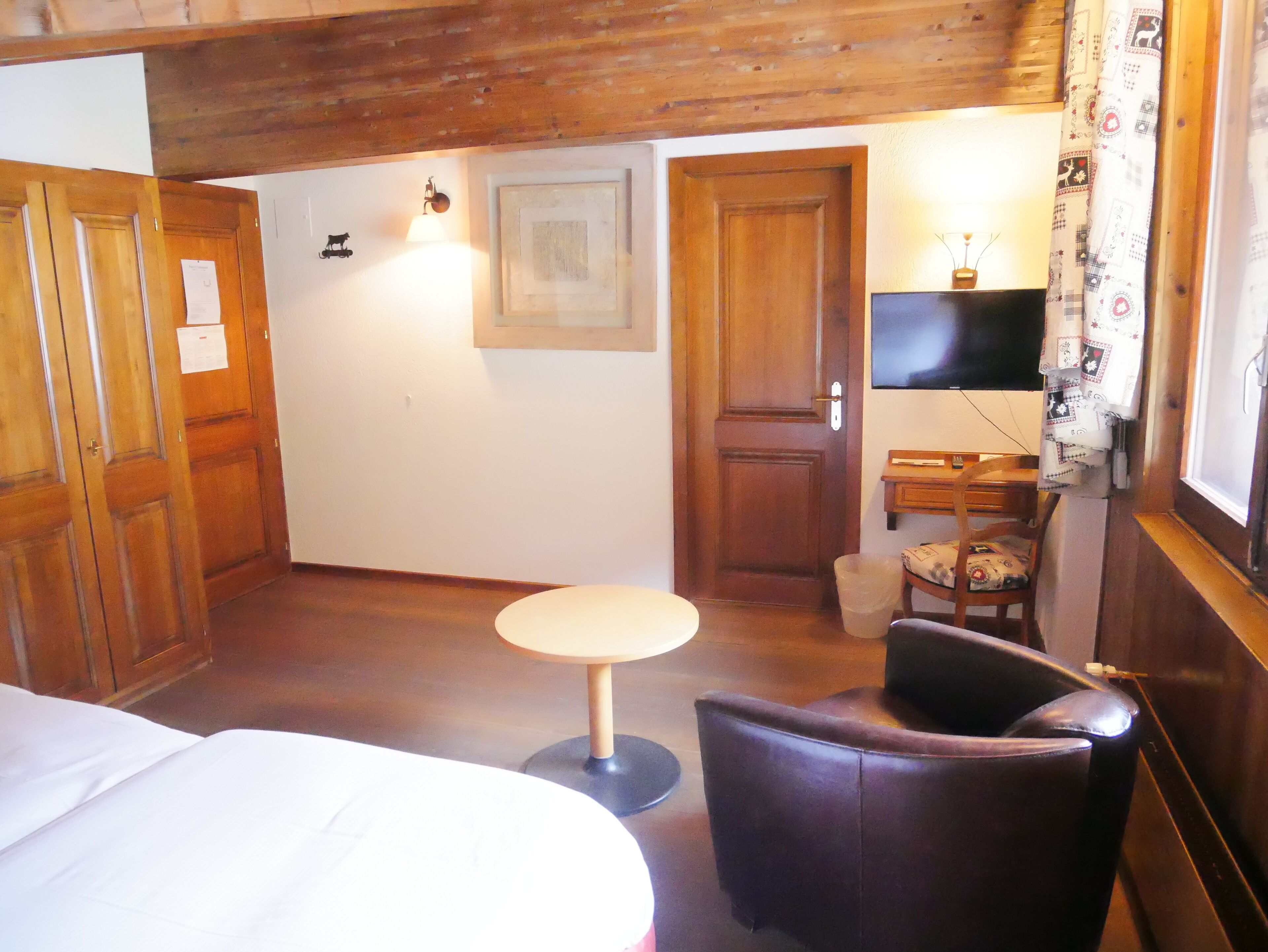 A room with a bed, a small round table, a leather chair, a wooden door, a wall-mounted TV, and a window with patterned curtains.