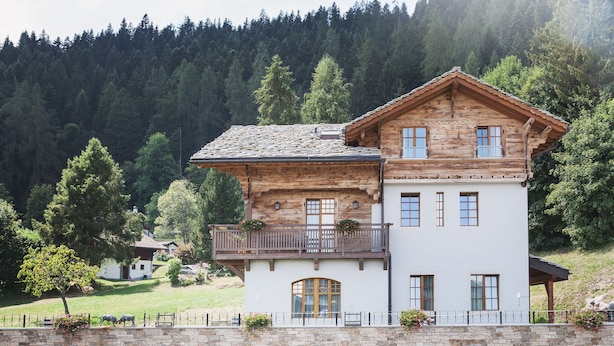 A two-story wooden house with a balcony, surrounded by a stone wall and a fence. - Between Boutique Hotel (Chamoson)