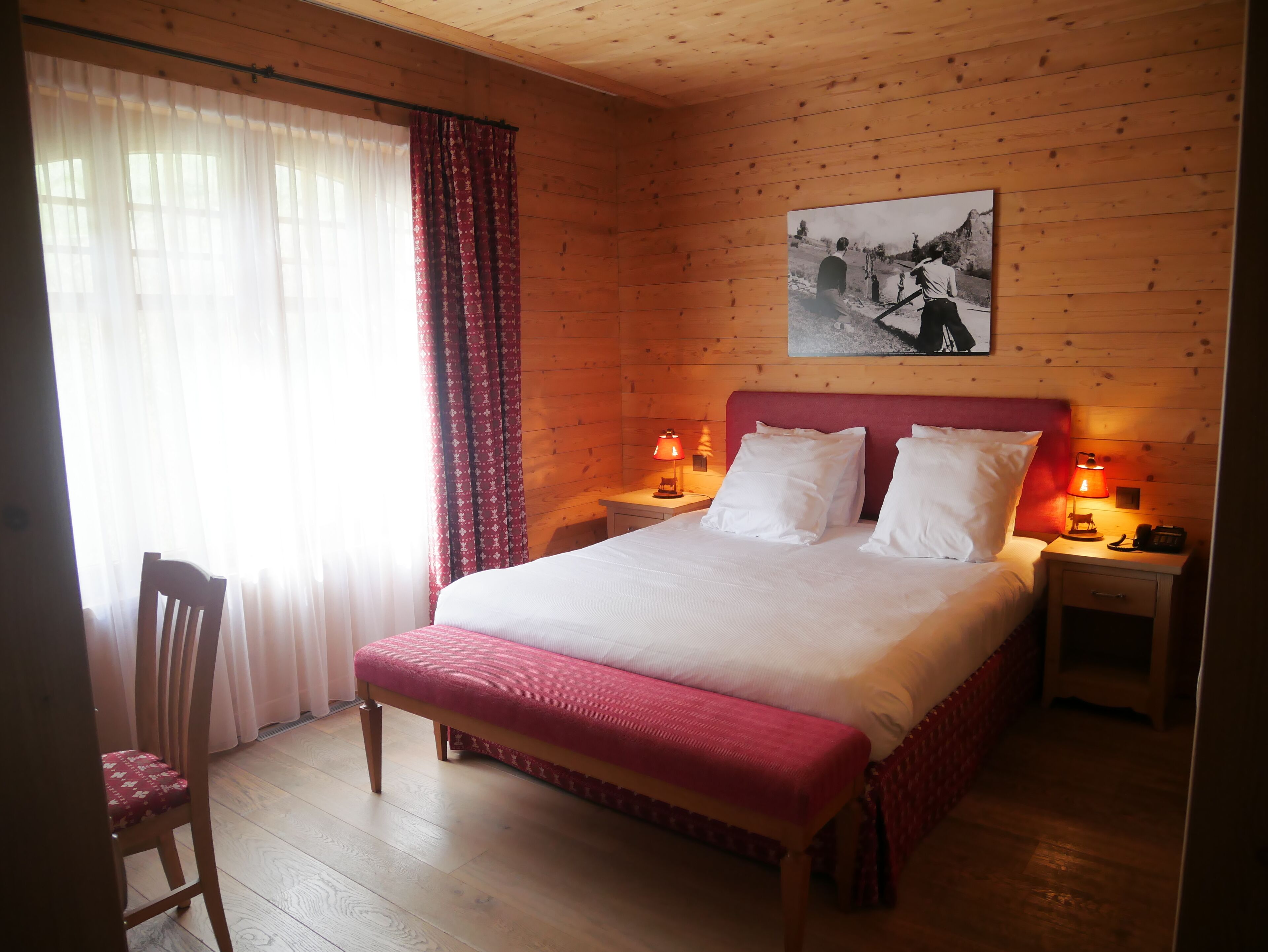 Una habitación con paneles de madera, una cama, dos mesitas de noche con lámparas, una silla y una ventana con cortinas.