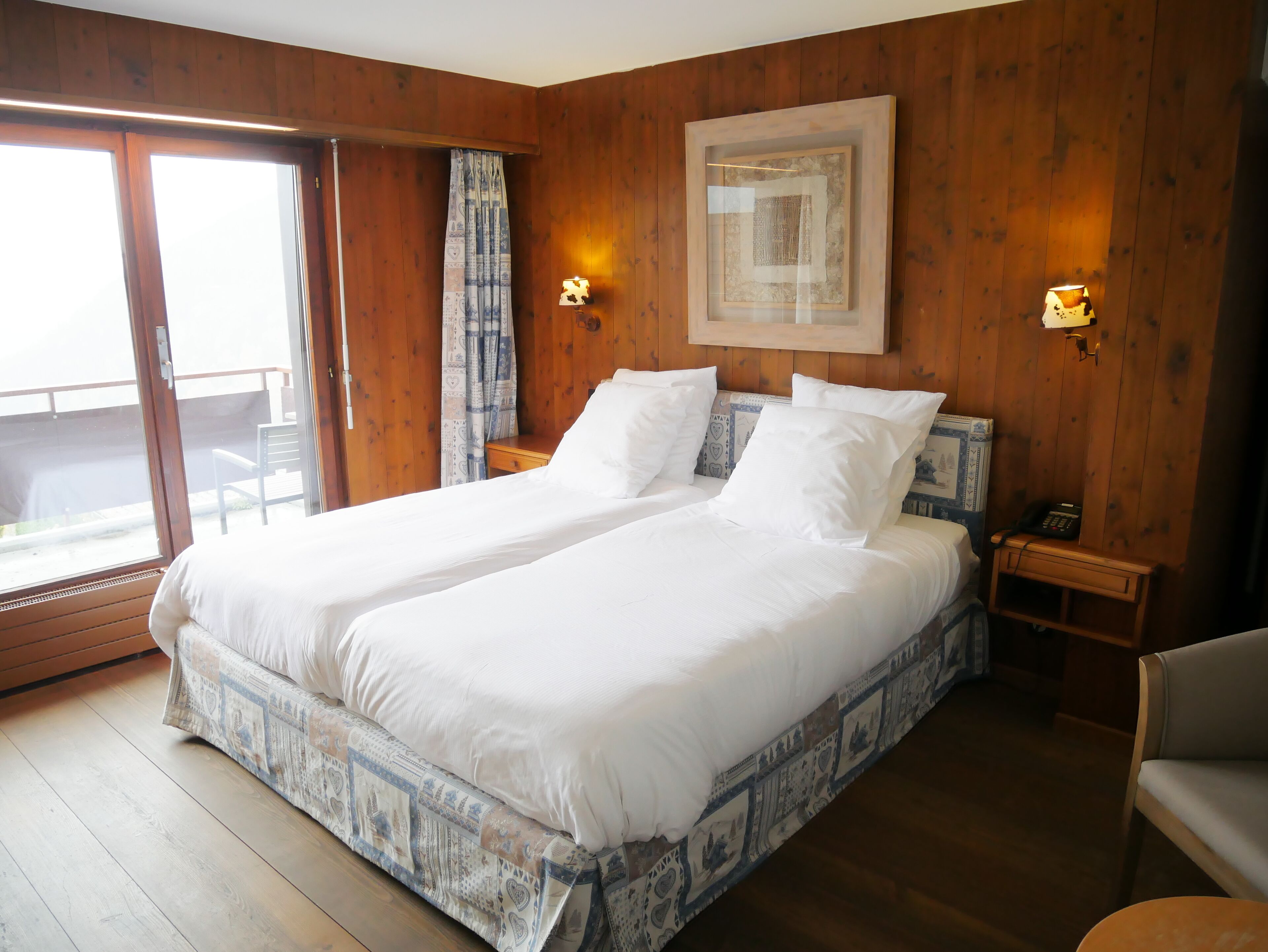 A double bed with white linens in a room with wooden walls and a large window.