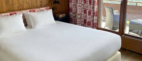 Un grand lit recouvert de draps blancs, dans une pièce aux murs en bois, donnant sur un balcon aménagé avec du mobilier d’extérieur.
