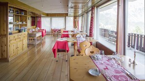 A wooden-floored dining area with tables set for dining, a kitchen area with wooden cabinets, and large windows offering a view of the outdoors. - Between Boutique Hotel (Chamoson)