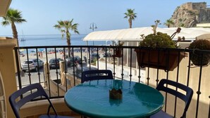 Un balcón con una mesa redonda y dos sillas, con vista a una playa donde se ven coches aparcados y palmeras.