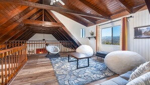 A spacious loft with a high ceiling, exposed wooden beams, a large window offering a mountain view, a cozy seating area with a sofa and armchairs, and a staircase leading to an upper level. - New!! Majestic Views, Pool, Sleeps 20+, 2 Mstr BR, close DT Yosemite, Bass Lake  (Oakhurst)