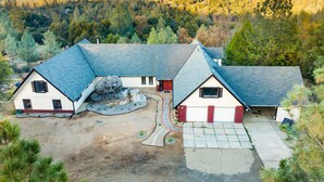 A two-story house with a large garage, surrounded by a forested landscape and mountains in the background. - New!! Majestic Views, Pool, Sleeps 20+, 2 Mstr BR, close DT Yosemite, Bass Lake  (Oakhurst)
