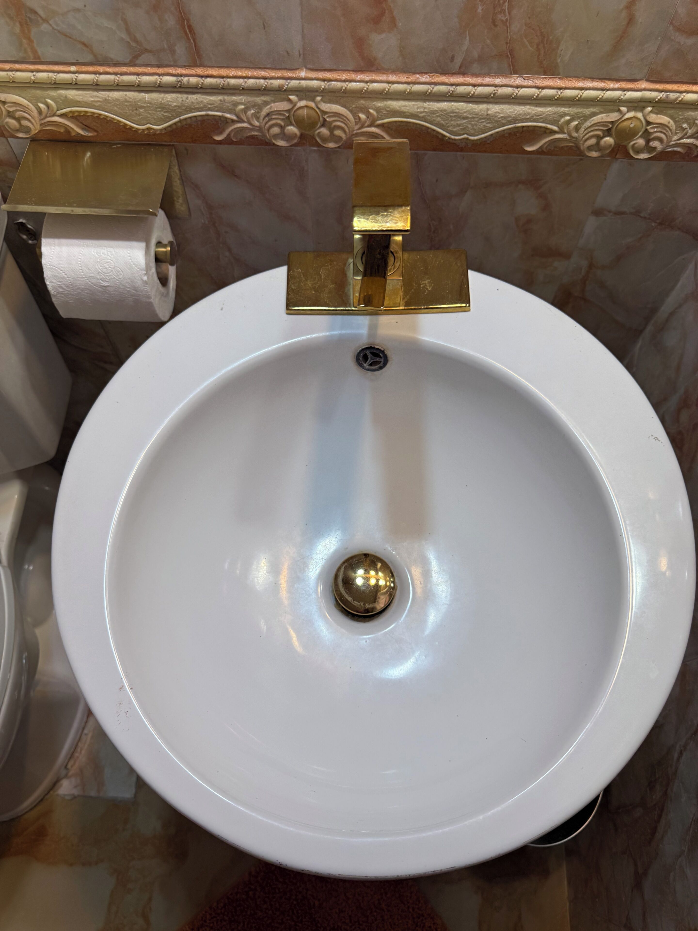 Un lavamanos blanco con grifo dorado, un dispensador de papel higiénico y una pared de mármol.
