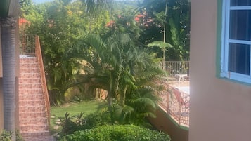 A view of a courtyard with a staircase leading to a balcony, surrounded by lush greenery and palm trees.