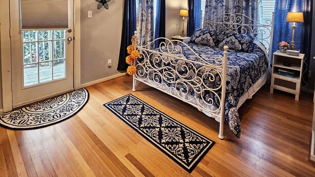 A bedroom with a bed, a ceiling fan, a window, curtains, a door, and a small nightstand with a lamp.