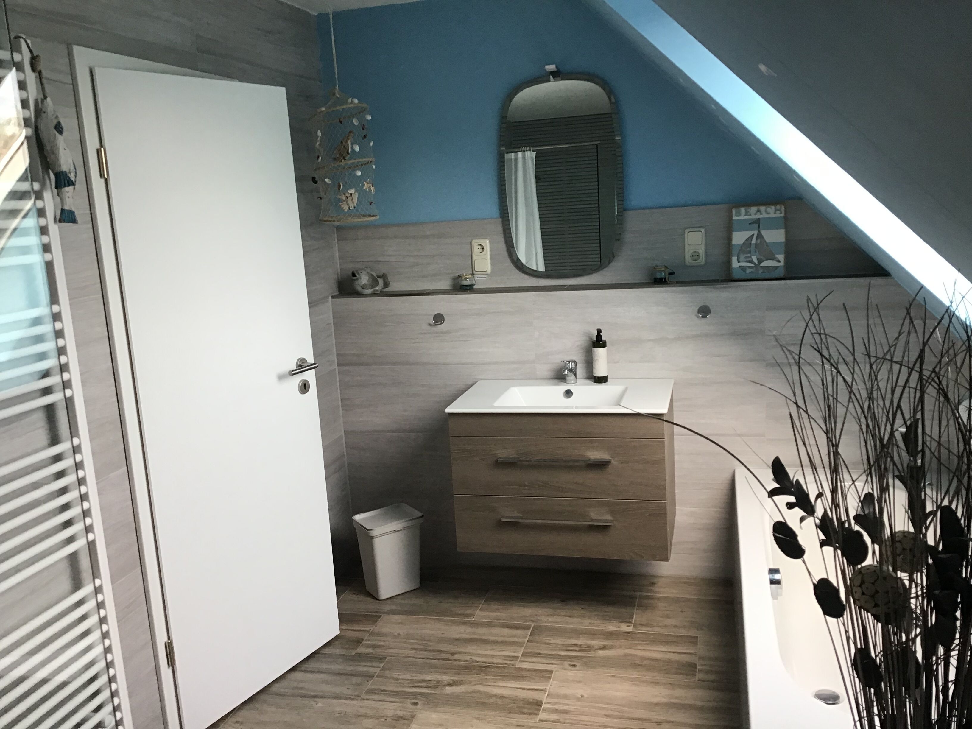 Un baño con un lavamanos blanco, un armario de madera y un espejo encima. Hay una puerta a la izquierda y un cubo de basura al lado del lavamanos.