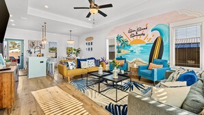 A modern living room with a large mural, a ceiling fan, and a kitchen area visible through an open door. - Heated Pool, Pickeball, Beach House (Panama City Beach)