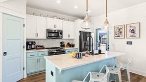 A modern kitchen with white cabinets, a central island, bar stools, and a hanging light fixture. - Heated Pool, Pickeball, Beach House (Panama City Beach)