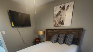 Une chambre à coucher avec un grand lit, un téléviseur à écran plat fixé au mur, une table de chevet avec une lampe et un tableau au-dessus du lit.