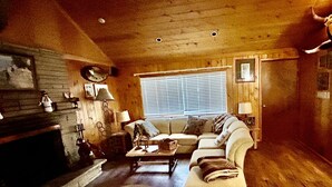 A cozy cabin interior with a fireplace, wooden walls, a sofa, and a window with blinds. - Cozy Utah Cabin (Kamas)