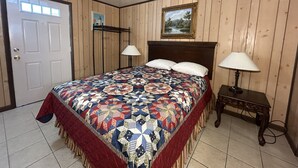 Quarto com parede revestida de madeira, cama com colcha estampada, duas luminárias de cabeceira, uma porta e um quadro na parede.