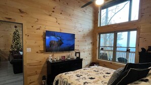 Un dormitorio de cabaña de troncos con cama, televisor, ventilador de techo y ventana con vistas a árboles.