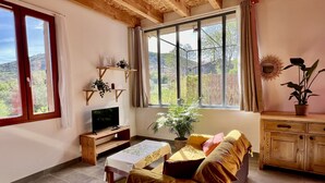 A cozy living room with a wooden ceiling, a sofa, a TV, a coffee table, and a view of a garden through large windows. - L'atelier du Salagou (Octon)
