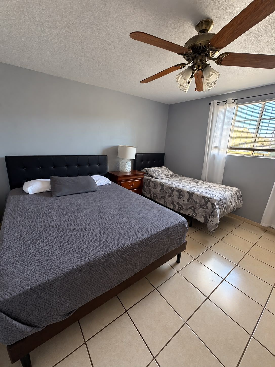 Une chambre avec un grand lit, un plus petit lit, une table de chevet avec une lampe, un ventilateur de plafond et une fenêtre avec des rideaux.