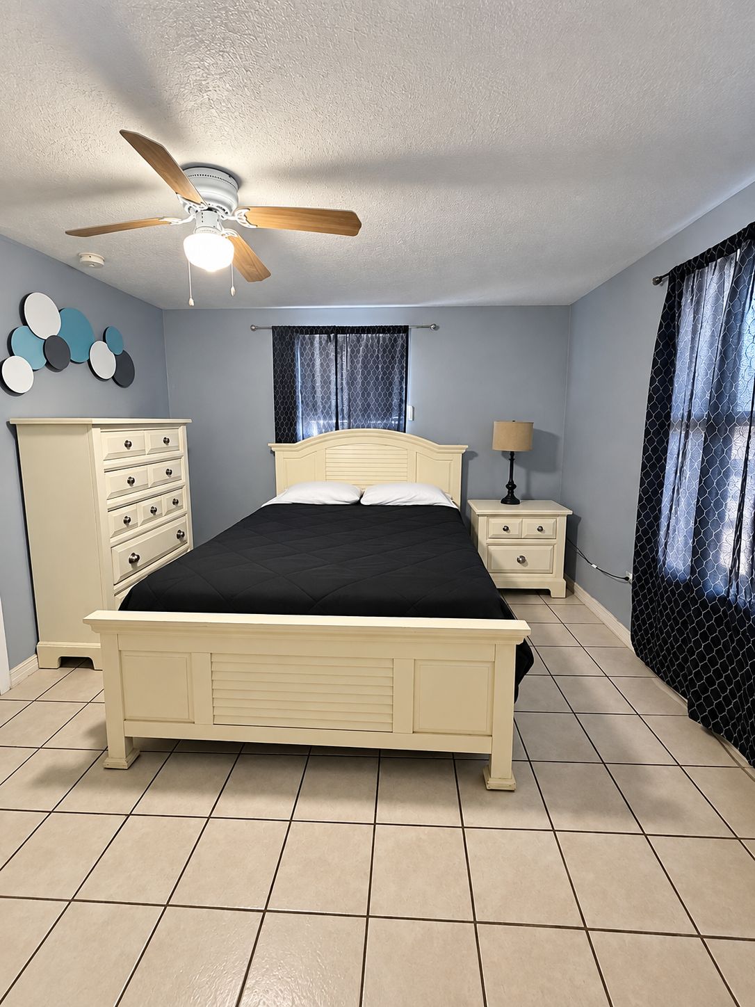A bedroom with a bed, a nightstand, a dresser, a ceiling fan, and a window with curtains.