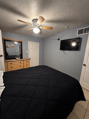 A bedroom with a bed, a ceiling fan, a TV mounted on the wall, a dresser, and a door.